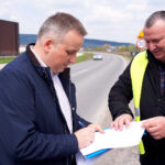 Two men on a highway shoulder review and sign documents, one in a bright safety vest.