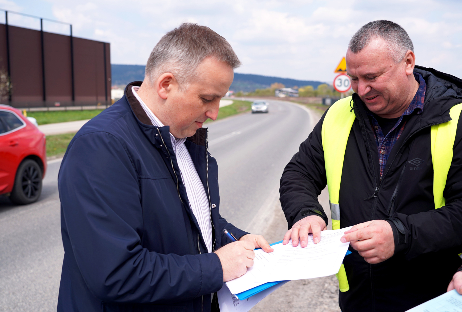Two men on a highway shoulder review and sign documents, one in a bright safety vest.