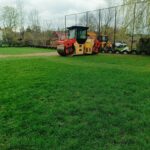 Yellow road roller and orange excavator parked on a grassy field beside a row of tires, with a chain-link fence and bare trees in the background.