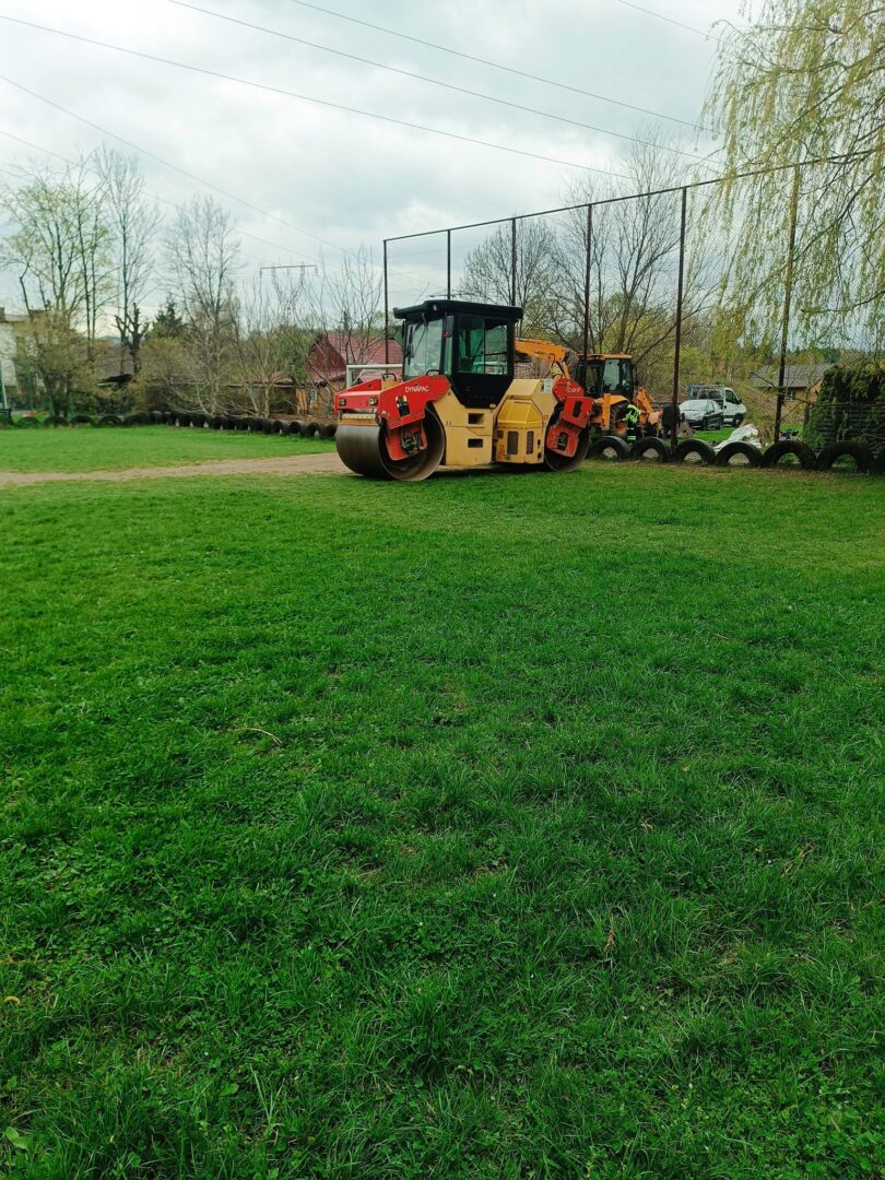 Yellow road roller and orange excavator parked on a grassy field beside a row of tires, with a chain-link fence and bare trees in the background.