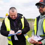 Two men in high-visibility vests by a rural road sign documents; one signs a clipboard while the other holds a pile of papers.