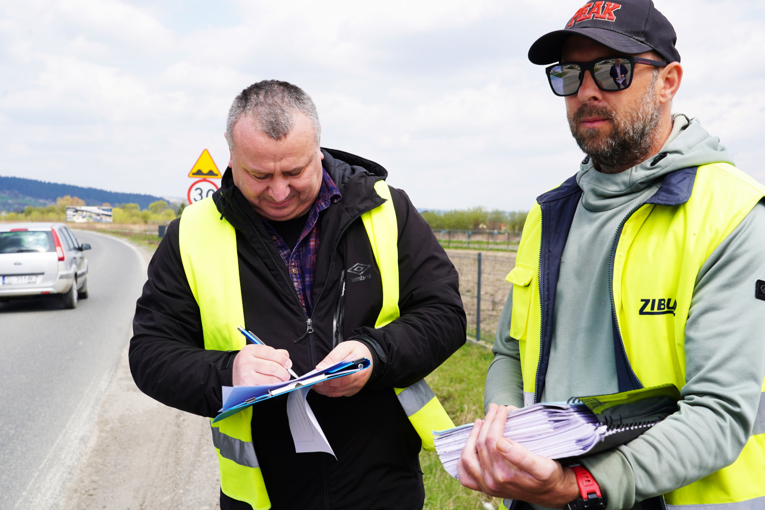 Two men in high-visibility vests by a rural road sign documents; one signs a clipboard while the other holds a pile of papers.