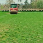 Red road roller on a grassy field with a row of tires and a chain-link fence in the background