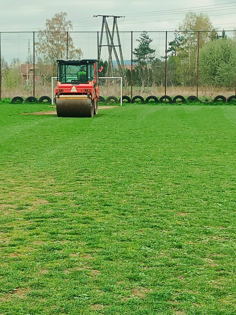 Red road roller on a grassy field with a row of tires and a chain-link fence in the background
