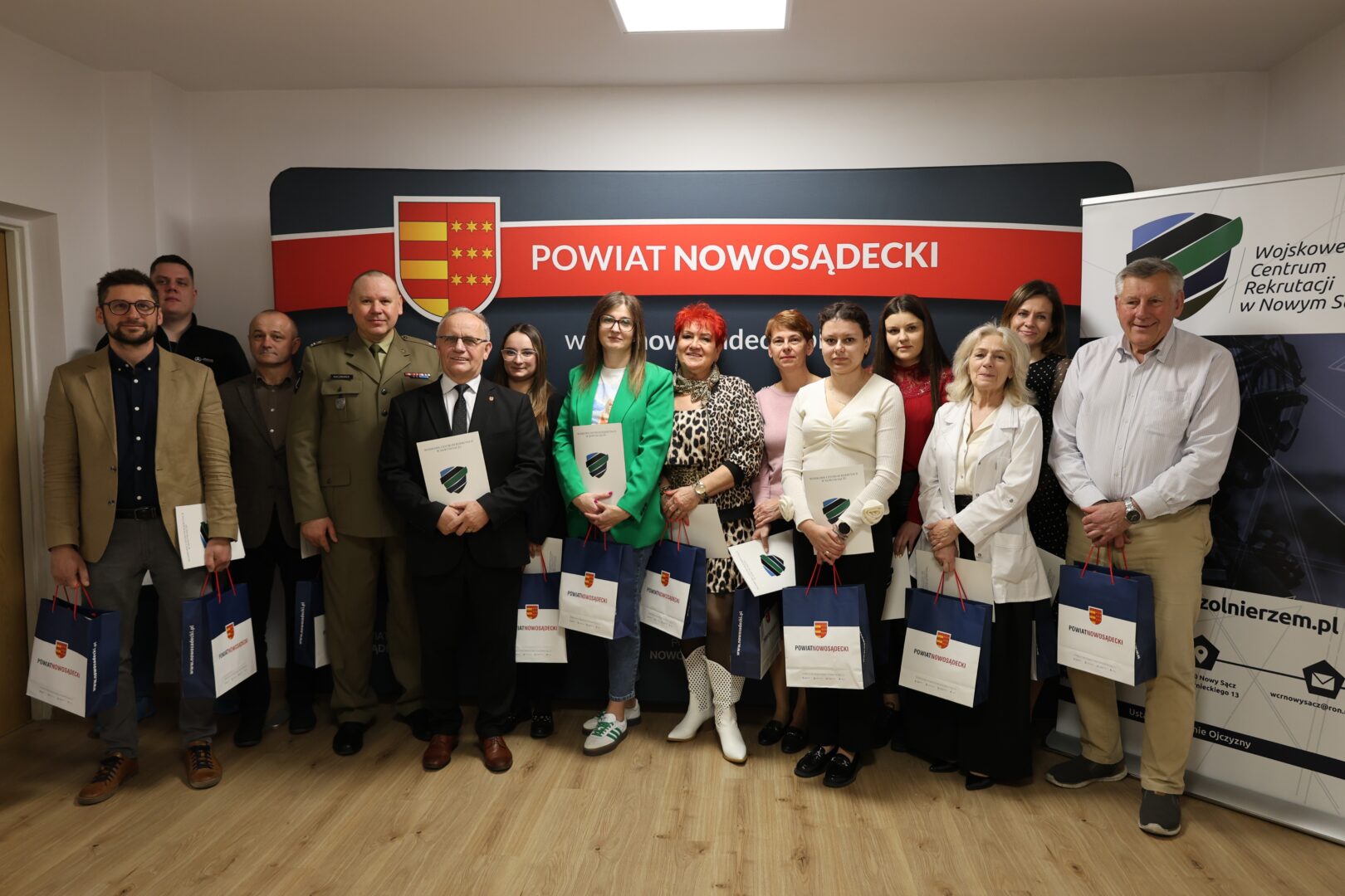 Group of men and women in formal attire posing with folders and gift bags during an awards event in front of a banner reading 'Powiat Nowosądecki'.