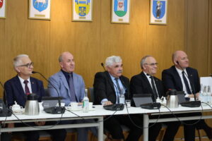 Five men in suits sit at a conference table with microphones during a formal panel discussion, wood-paneled walls behind them with crest portraits.