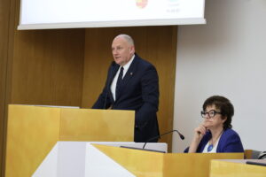 Man in a navy suit speaks into a microphone at a wooden podium, while a woman with glasses sits nearby, listening in a conference room.