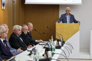 A speaker stands at a yellow podium addressing a panel of men seated at a long table with microphones in a conference room.