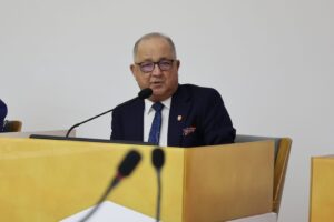 Older man in glasses, white shirt and navy suit speaking into a microphone at a wooden podium.