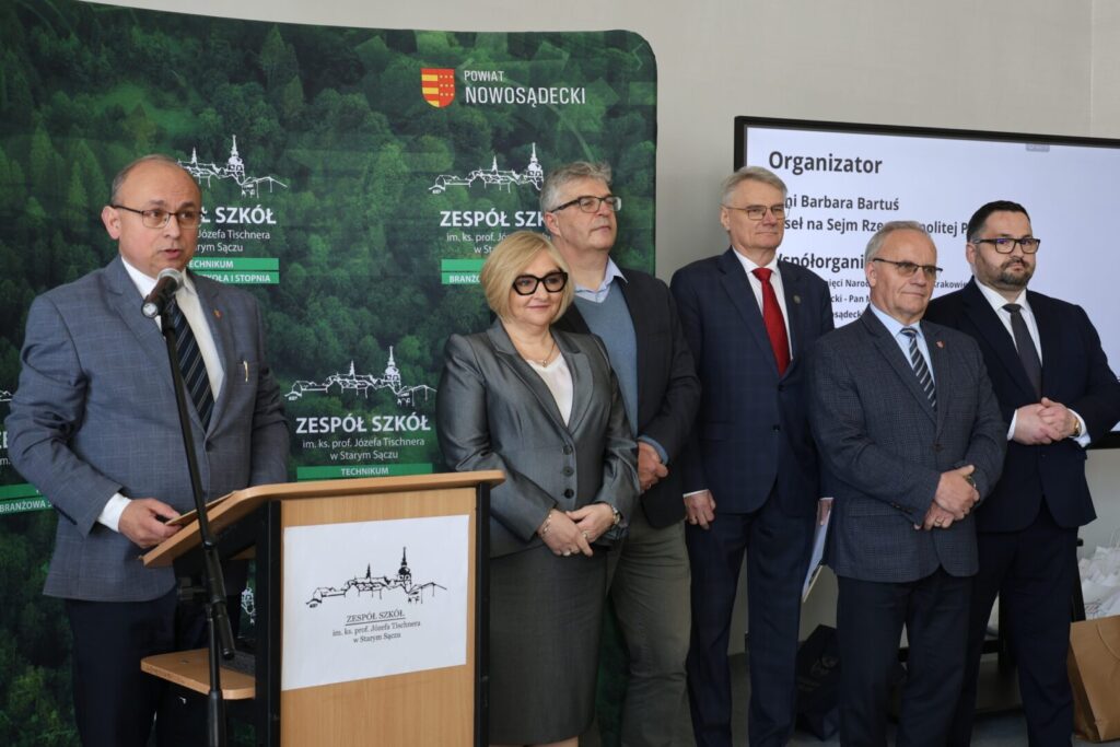 Six officials in suits stand at a podium during a formal event, with a forest-themed backdrop and a screen labeled Organizer in the background.