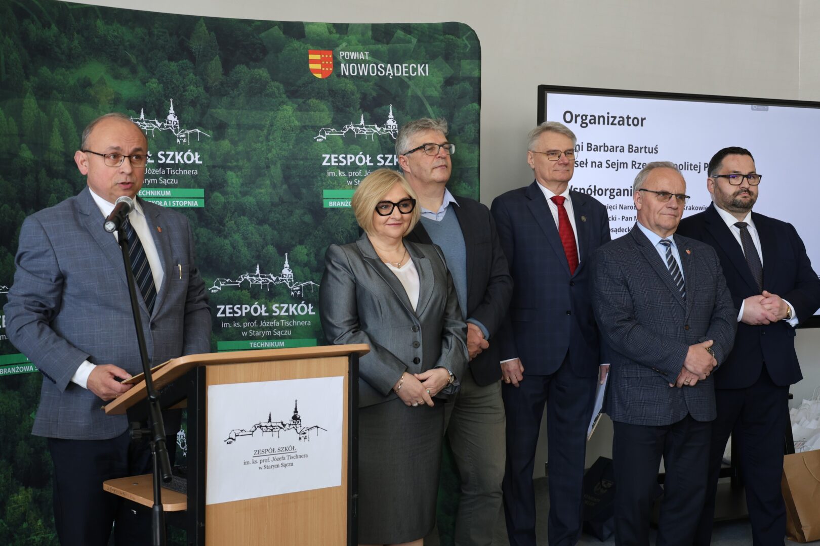Six officials in suits stand at a podium during a formal event, with a forest-themed backdrop and a screen labeled Organizer in the background.