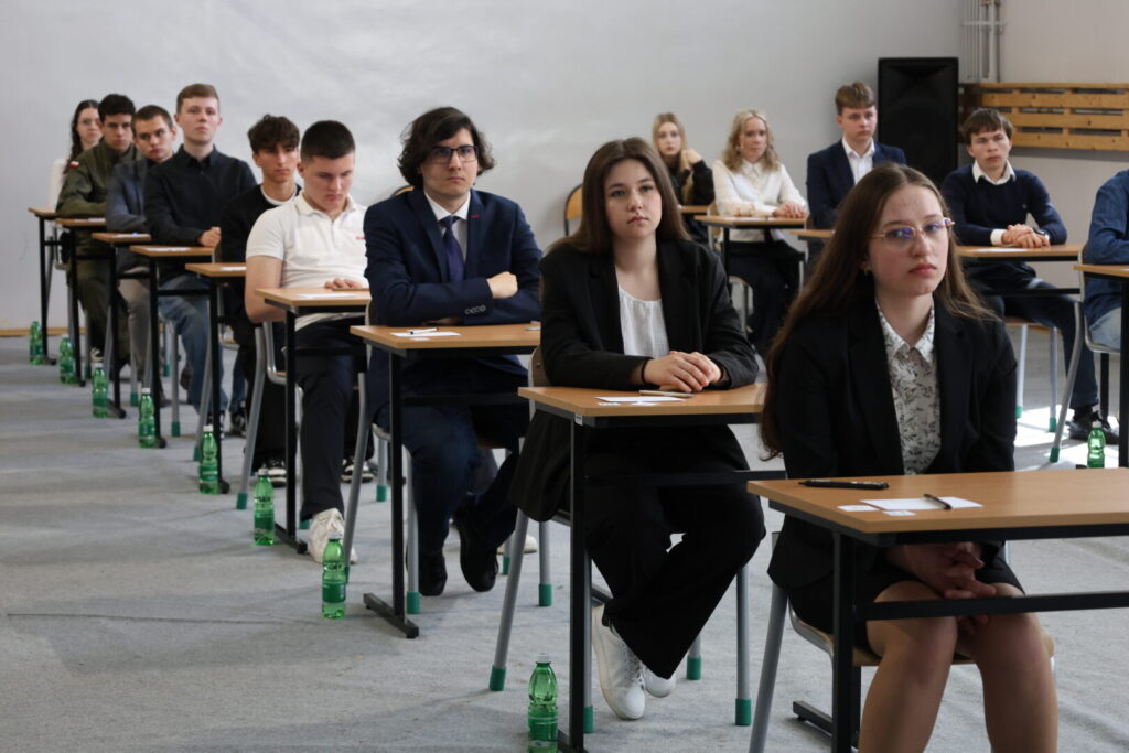 Classroom exam scene with students seated at desks; two girls in front wearing blazers, rows of peers behind, water bottles along the aisle.