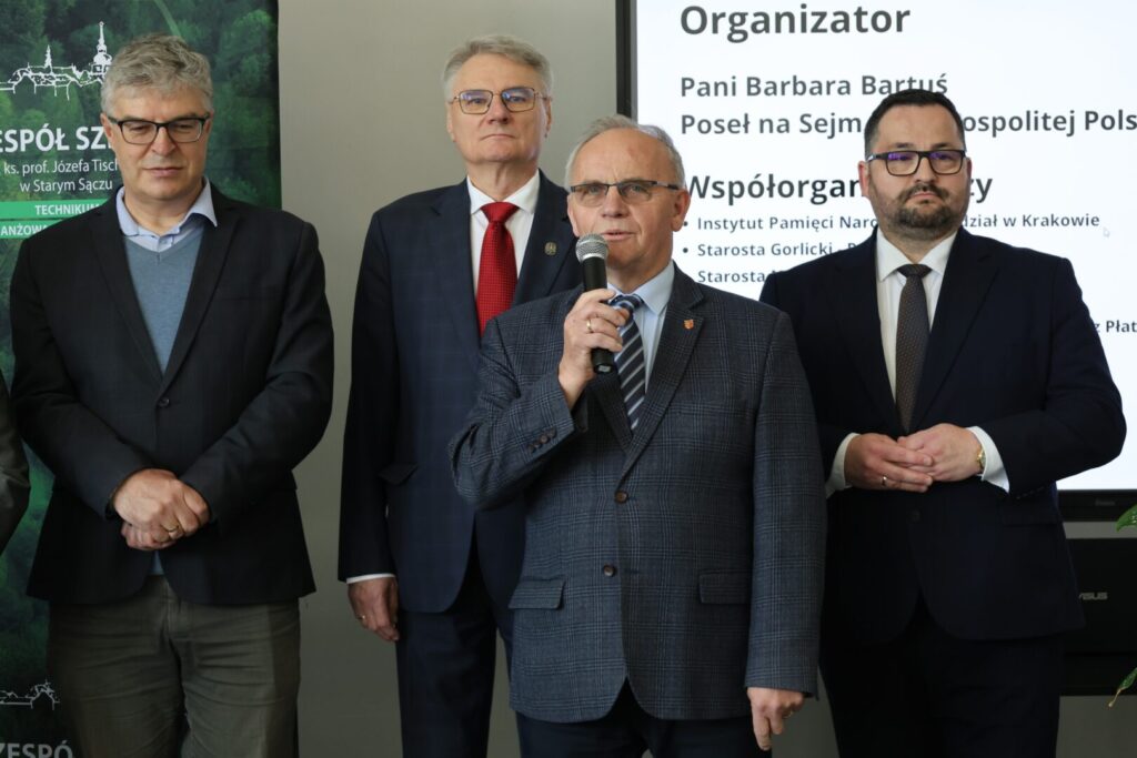 Four men in business suits stand at a formal event; the man in the center-right speaks into a microphone while others listen.