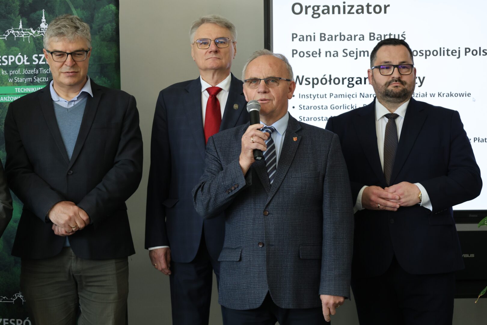 Four men in business suits stand at a formal event; the man in the center-right speaks into a microphone while others listen.