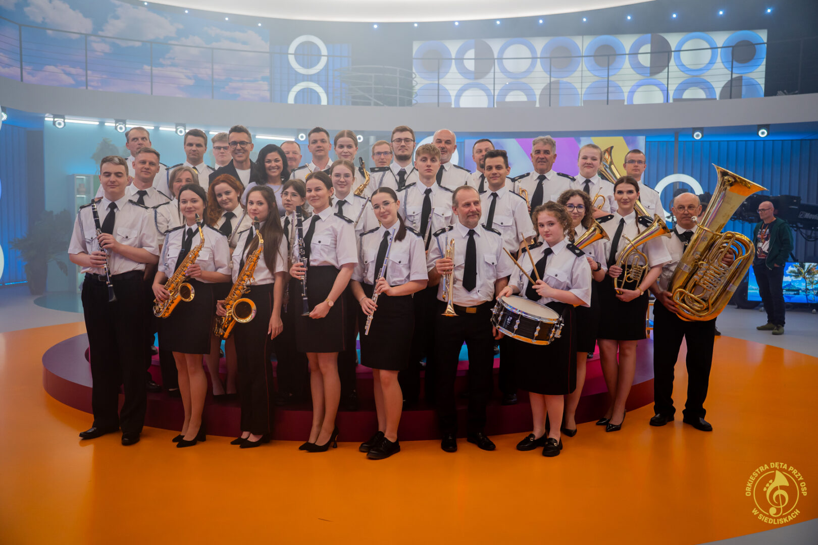 Large ensemble of uniformed musicians posing with brass and woodwind instruments on a bright studio stage.