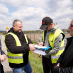Four men outdoors by a rural road, two wearing high-visibility vests, signing papers from a clipboard with others watching.