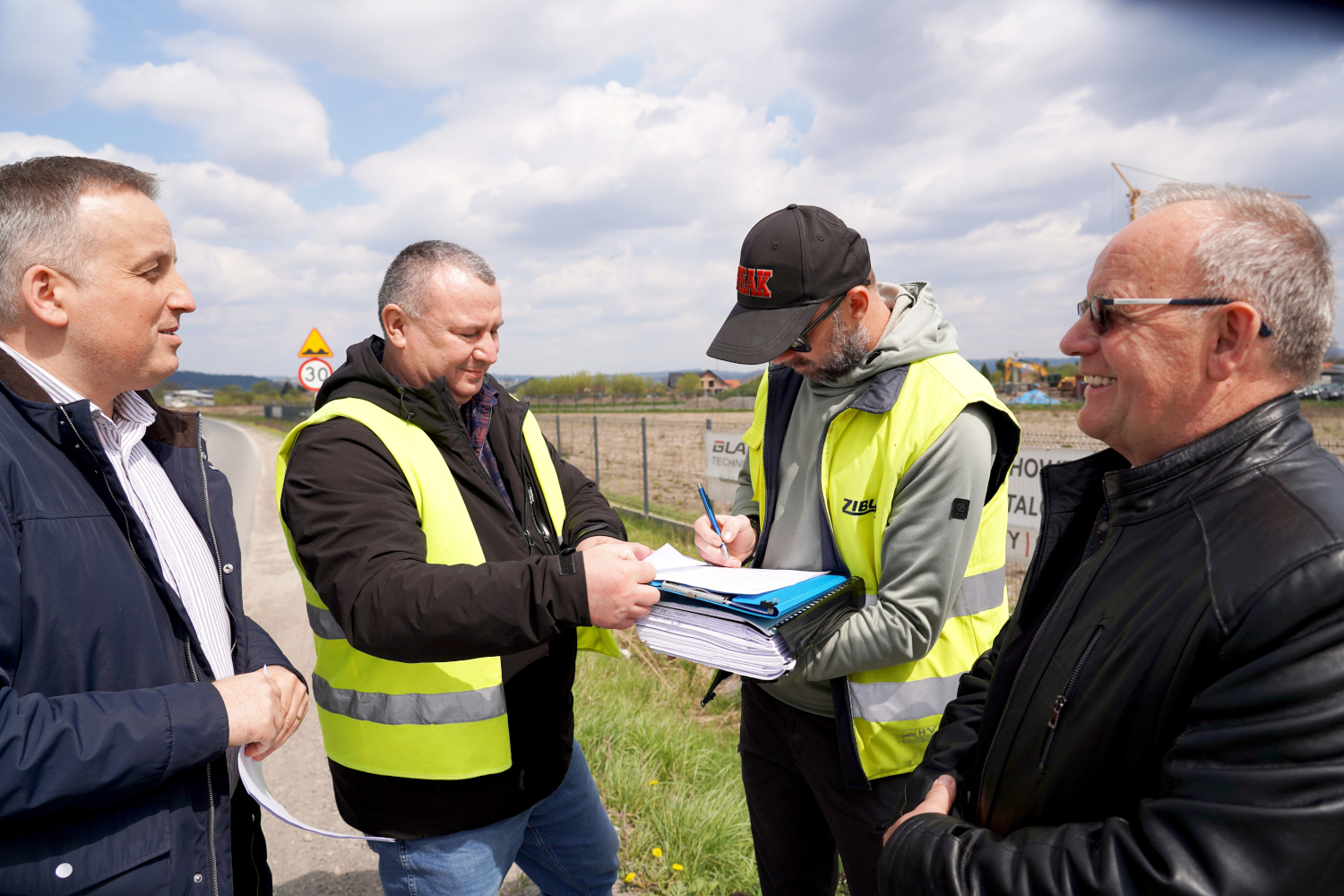 Four men outdoors by a rural road, two wearing high-visibility vests, signing papers from a clipboard with others watching.