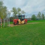 Red-and-beige road roller parked on a grassy field beside a small soccer goal under a cloudy sky.