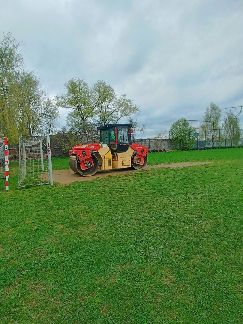 Red-and-beige road roller parked on a grassy field beside a small soccer goal under a cloudy sky.