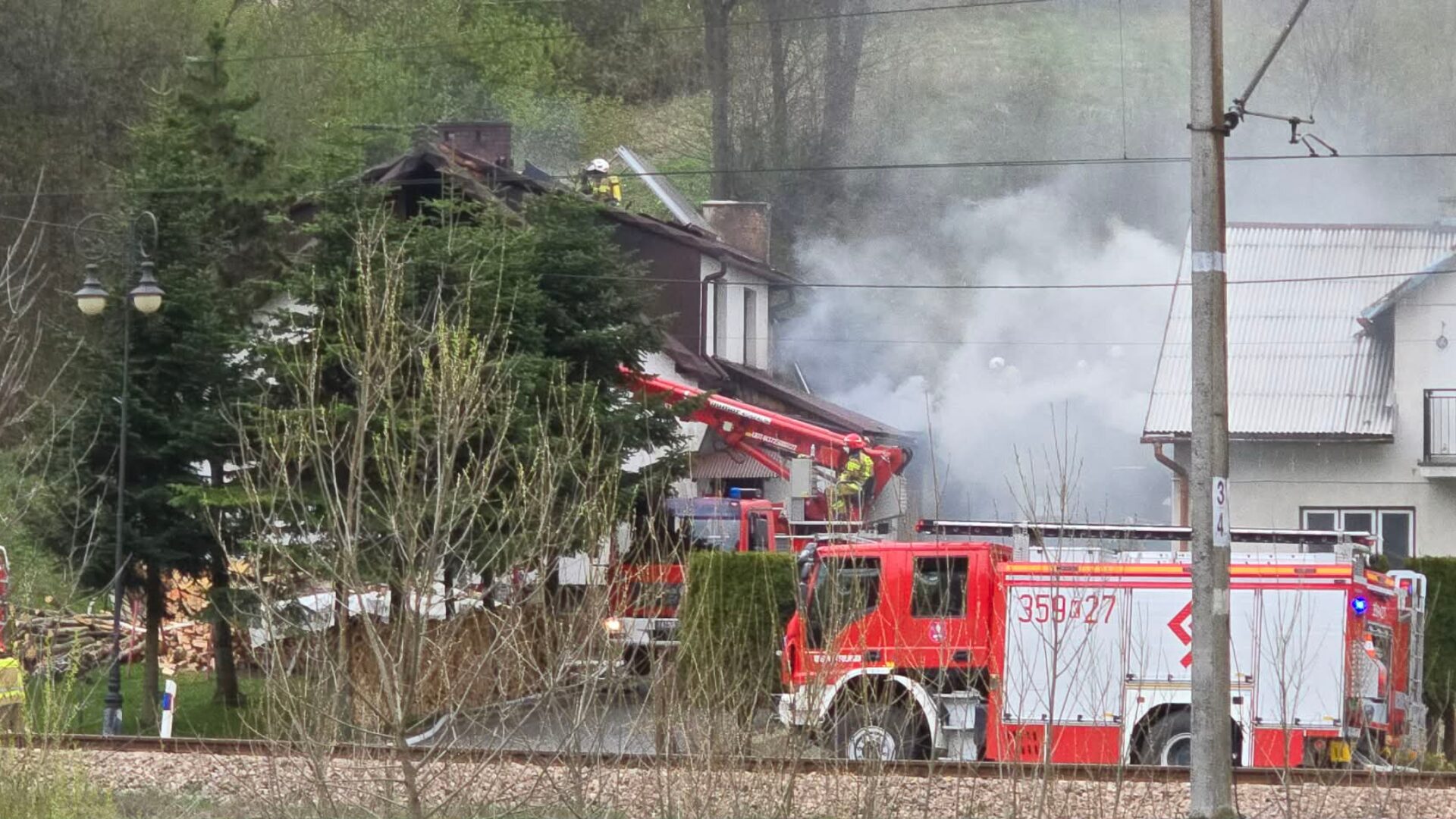 Firefighters on a ladder perched against a burning house with thick smoke, while red fire engines park nearby in a residential area.