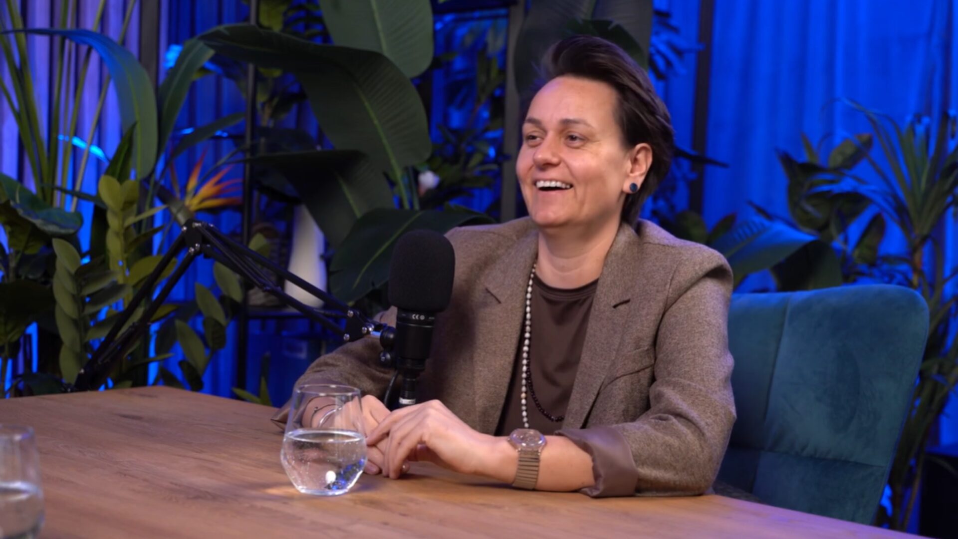 Smiling woman in a brown blazer sits at a wooden table with a microphone in front of her during a podcast interview, a glass of water beside her, blue studio lighting and tropical plants in the background.