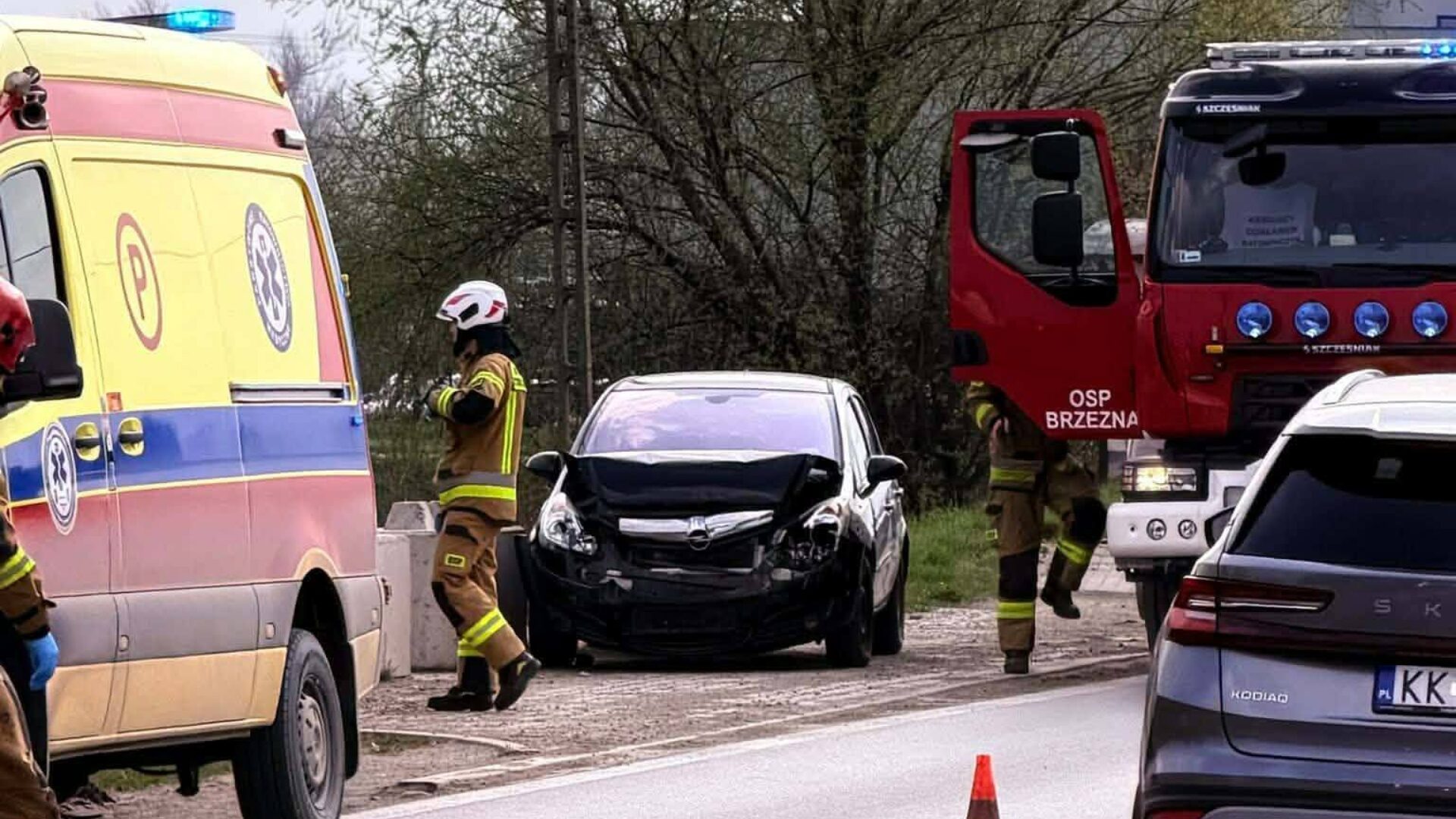 Emergency responders in protective gear attend a vehicle crash: a black car with front-end damage sits on a roadside as an ambulance and a red fire truck block the scene.