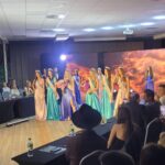 Beauty pageant stage: contestants in colorful gowns with sashes and crowns, holding flowers as audience watches from tables on either side.