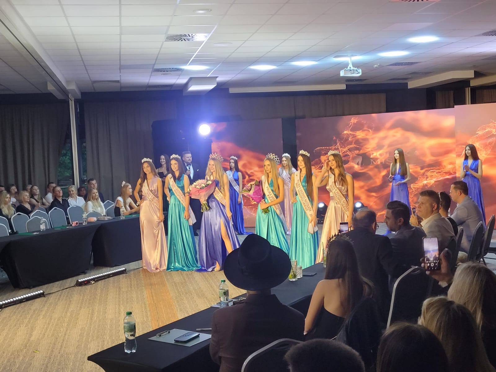 Beauty pageant stage: contestants in colorful gowns with sashes and crowns, holding flowers as audience watches from tables on either side.