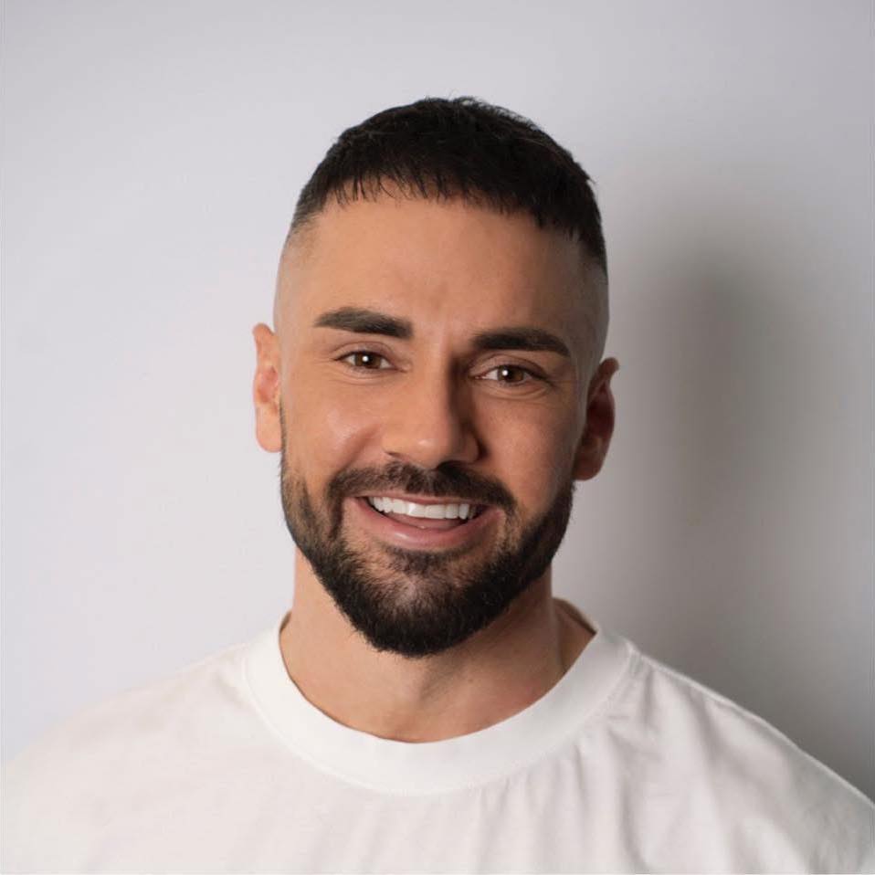 Smiling man with short dark hair and trimmed beard wearing a white shirt against a light gray background.