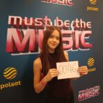 Young woman with long brown hair posing in front of a 'Must Be the Music' backdrop, holding a card that reads 6046.