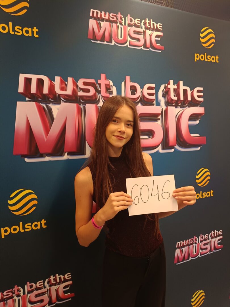 Young woman with long brown hair posing in front of a 'Must Be the Music' backdrop, holding a card that reads 6046.