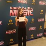 Young girl posing for a photo at a music event, holding a sign that reads 6046 in front of a 'Must Be The Music' backdrop with Polsat logos.