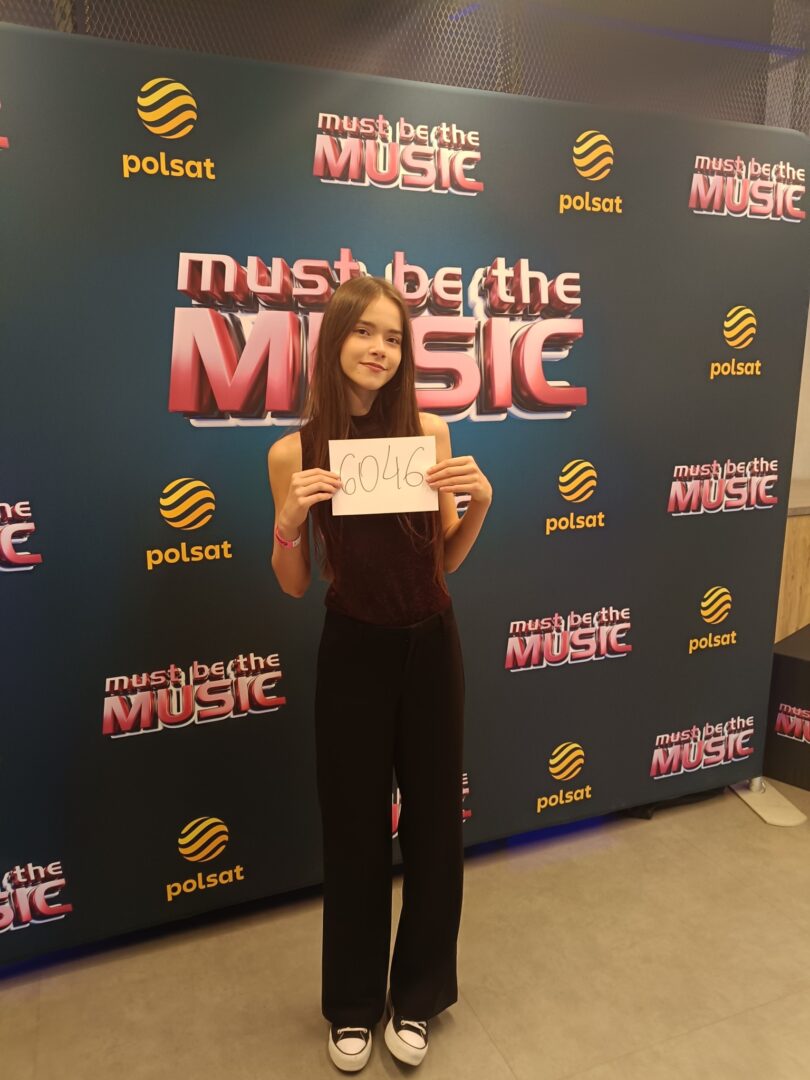 Young girl posing for a photo at a music event, holding a sign that reads 6046 in front of a 'Must Be The Music' backdrop with Polsat logos.