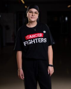Young person wearing a black headscarf and glasses, in a black t‑shirt that reads 'CANCER FIGHTERS', standing in a dim indoor space.
