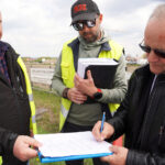 Three men in high-visibility jackets sign or review documents at an outdoor work site.