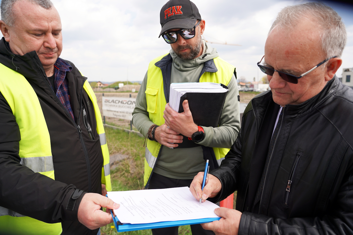 Three men in high-visibility jackets sign or review documents at an outdoor work site.