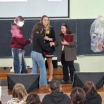 Student on stage shakes hands with a presenter, while another woman holds a microphone in a classroom ceremony setting.