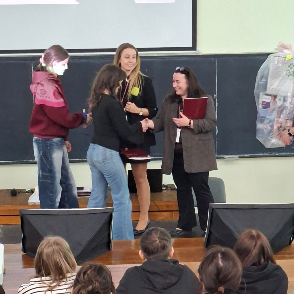 Student on stage shakes hands with a presenter, while another woman holds a microphone in a classroom ceremony setting.