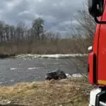 Fire truck on the riverbank beside a car stranded in the water, trees in the background.