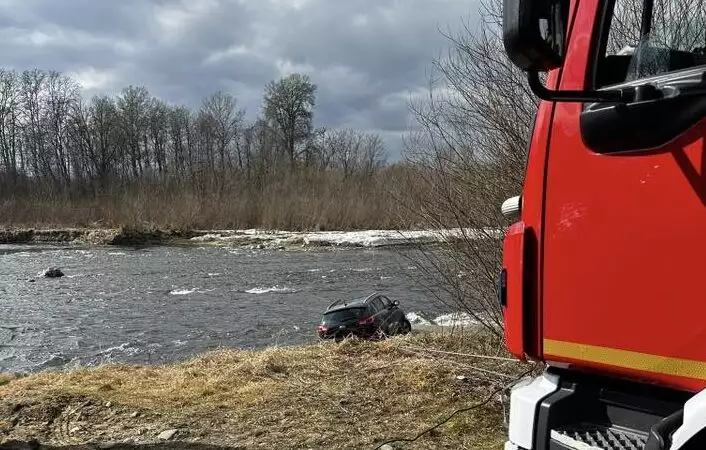 Fire truck on the riverbank beside a car stranded in the water, trees in the background.