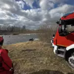 Firefighters in red gear by a riverbank, with a red rescue truck on the right; a car is submerged in the water.
