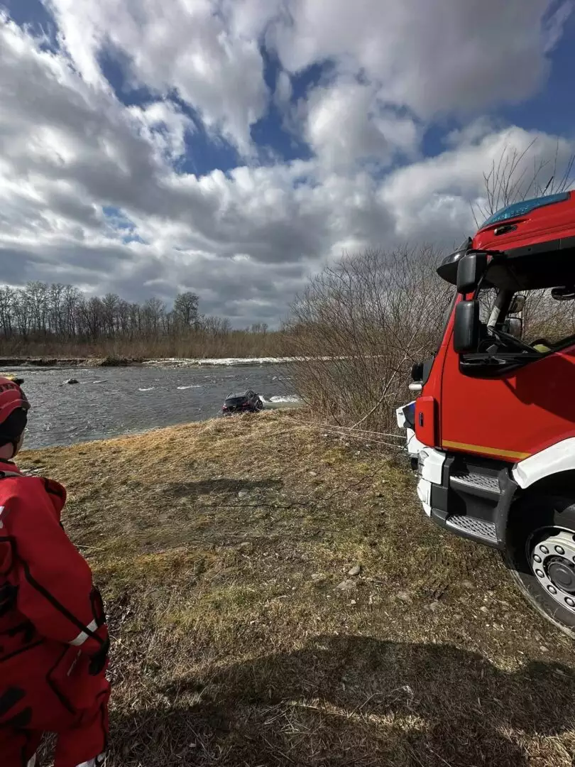 Firefighters in red gear by a riverbank, with a red rescue truck on the right; a car is submerged in the water.