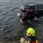 A black SUV is partially submerged in a river; rescue divers in red and yellow gear are securing it with ropes from the water and shore.