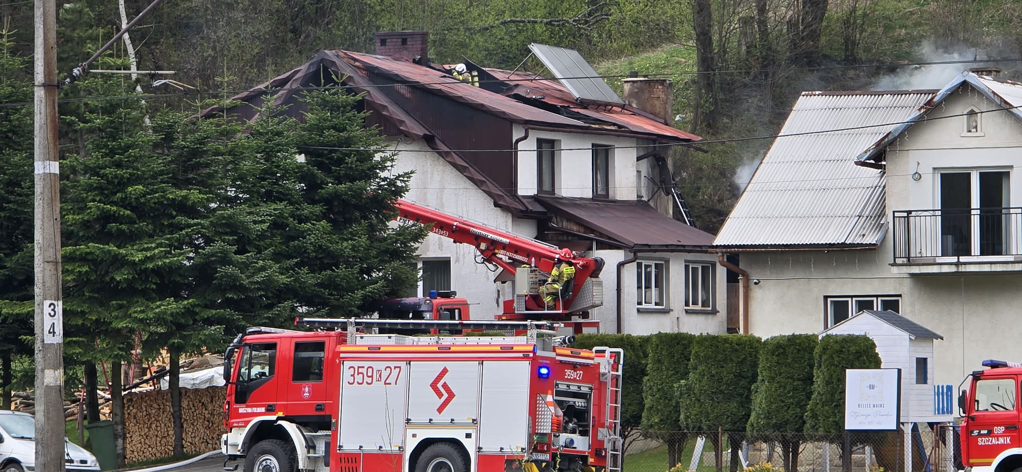 Firefighters on a pitched red roof with a ladder truck below and smoke in the background; a red fire engine is parked in front of a white house district scene.