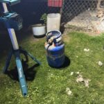 Blue propane gas cylinder on a grassy yard next to a blue Makita work stand; a chain-link fence and a dog visible in the background.