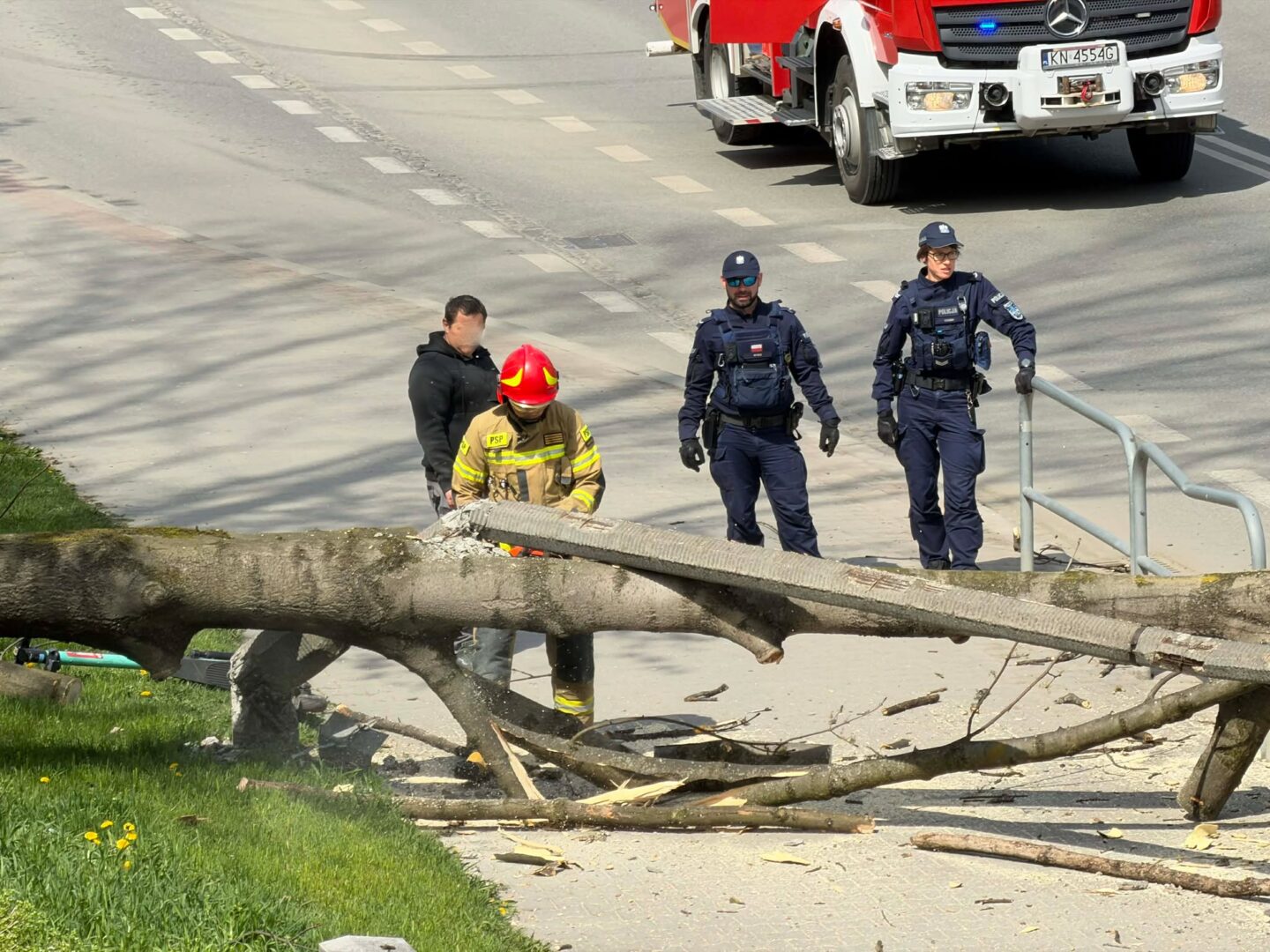 Firefighter in yellow gear and red helmet removing a fallen tree blocking a sidewalk while two police officers stand nearby; truck in background.