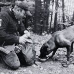 Hiker in a cap and jacket kneels beside a large backpack in a forest, as his dog drinks from a bowl on the ground nearby.