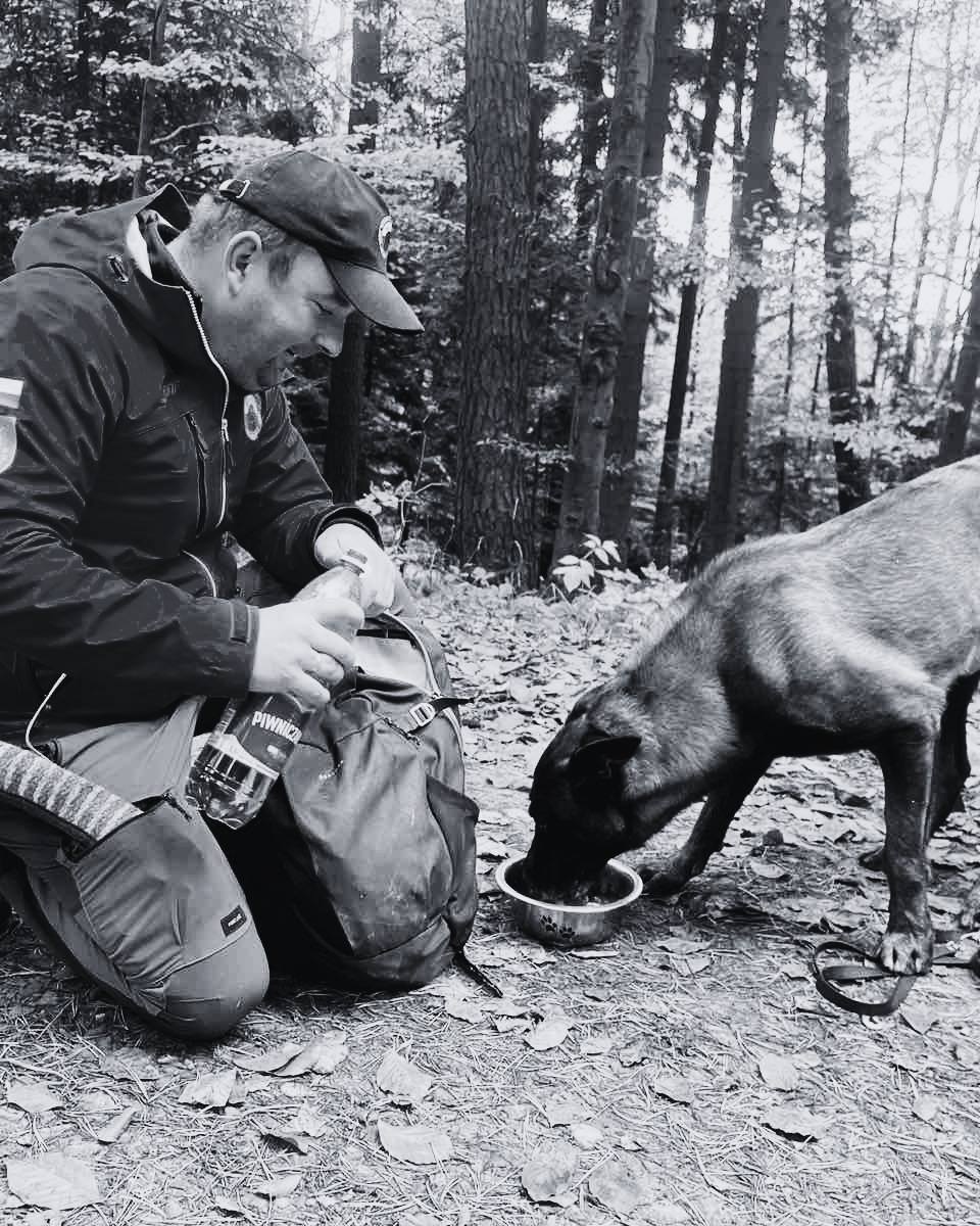 Hiker in a cap and jacket kneels beside a large backpack in a forest, as his dog drinks from a bowl on the ground nearby.