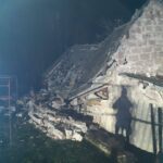 Ruined stone building with a collapsed roof and scattered bricks at night, with a long shadow of a person in the foreground.