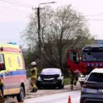 Emergency responders at a car crash on a road, with a damaged black sedan, an ambulance, and a fire truck nearby.